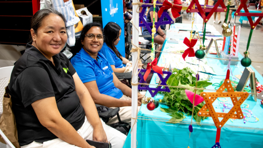 Three STEPS AMEP clients sit at their Christmas stall in Bunnings Palmerston. The table is covered in a bright blue tablecloth, with several goods available for sale spread over it.