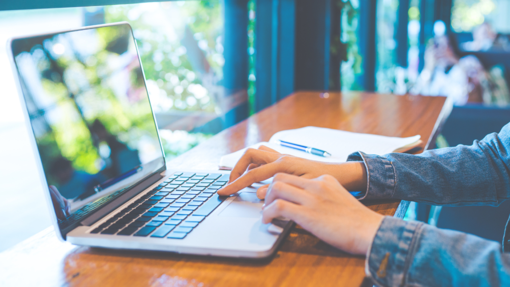 Close-up of hands typing on a laptop at a wooden desk with an open notebook and pen nearby, in a bright workspace.