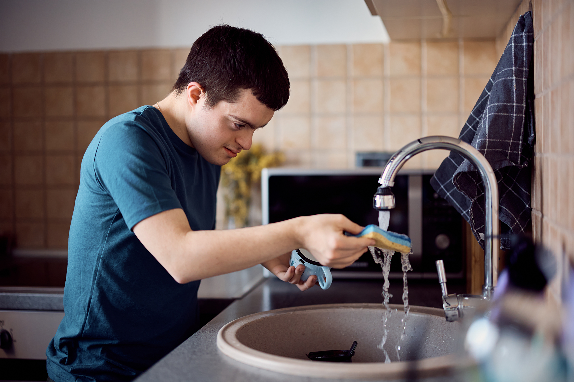 A person with a disability is washing a dish under running water at a kitchen sink.