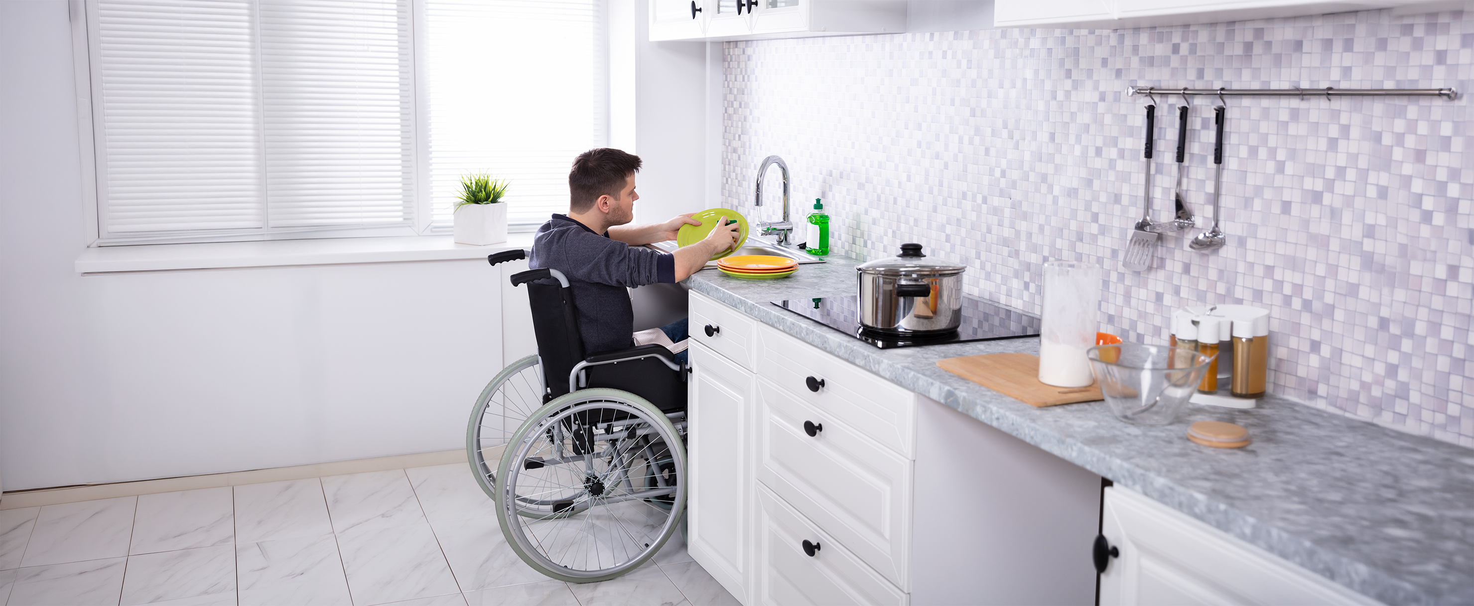 A person using a wheelchair is washing dishes at a kitchen sink, with accessible counter space and utensils arranged within easy reach.