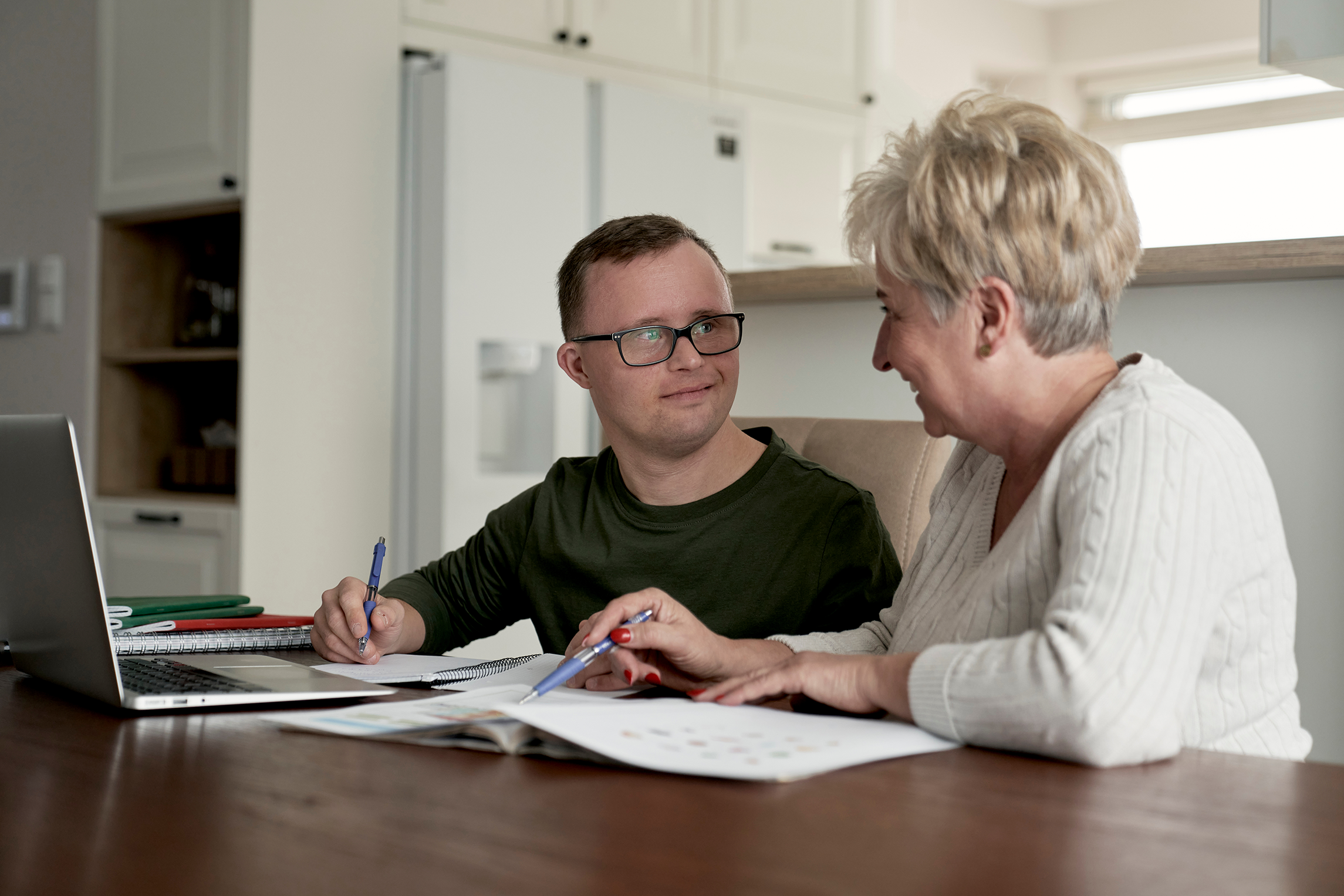 A person supports another person at a kitchen table, working together through written materials and notes beside an open laptop. The scene reflects an Individualised Living Option (ILO) arrangement, showing collaborative planning and day‑to‑day assistance in a home environment.