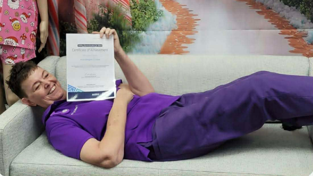 Anna lies on a lounge, holding her certificate of completion and smiling at someone off camera. She is wearing purple scrubs.