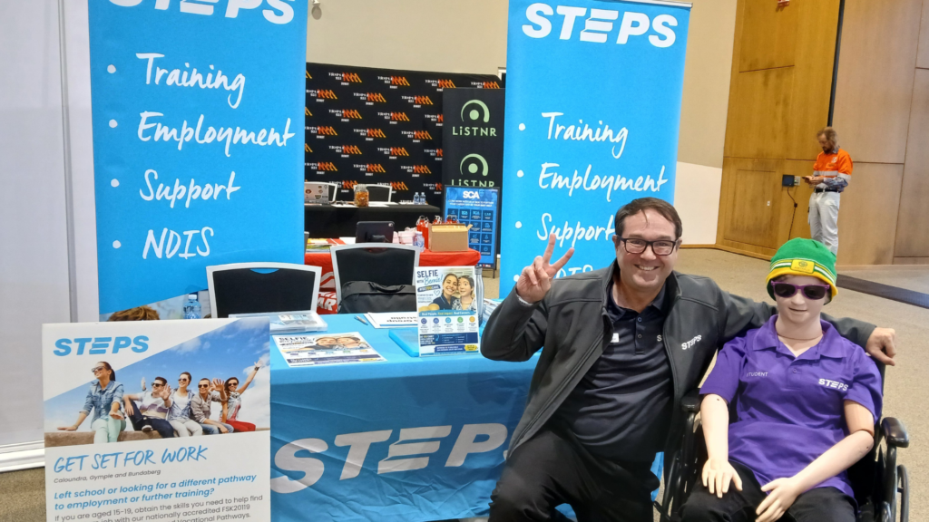 A man poses with a dressed mannequin in a wheelchair. They are in front of a blue stall with STEPS signage.