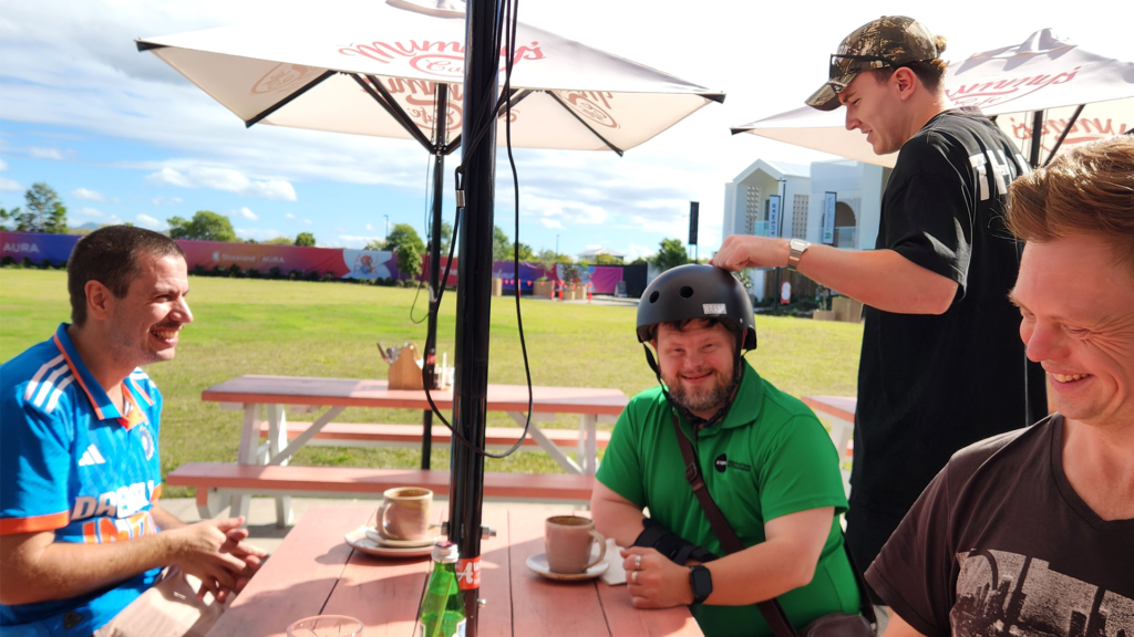 A candid photo of three men sitting at an outdoor cafe table on a sunny day, smiling and laughing. A fourth man is standing and smiling with them.