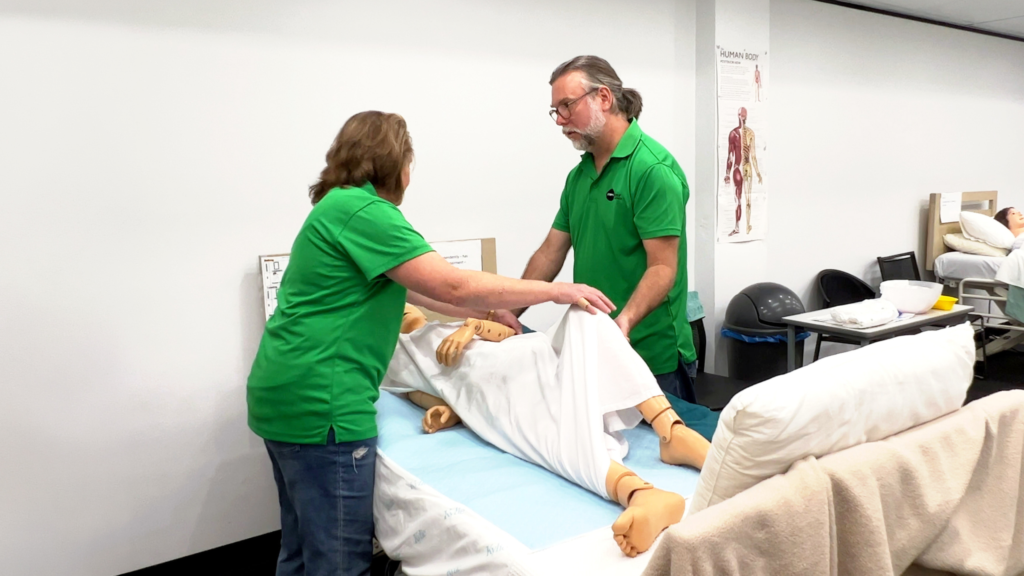Two participants in STEPS shirts practise a patient‑handling technique with a medical training mannequin in a simulated care environment.