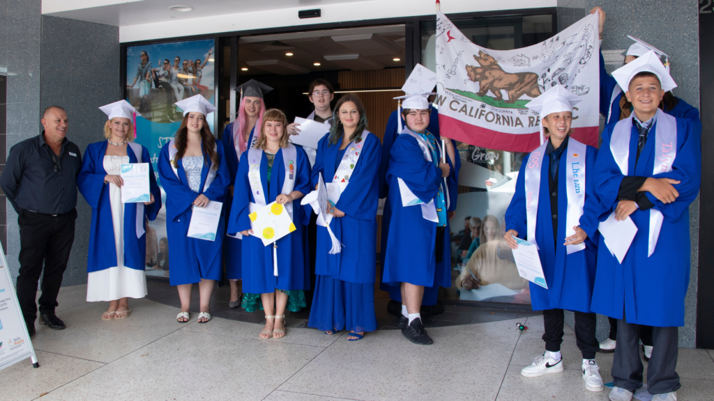 Eleven STEPS Get Set for Work graduates, all young adults, stand outside of the STEPS Bulcock Street site entrance, adorned in bright blue graduation gowns with white caps and sashes. They are joined by Michael Williamson, a member of the STEPS Executive Leadership team.