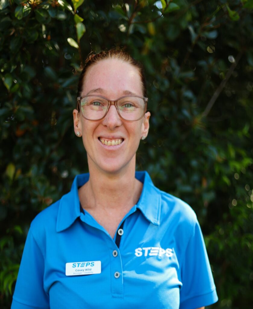 STEPS Disability Manager Casey smiles at the camera. She is wearing a bright blue STEPS polo shirt and is standing in front of green hedges.