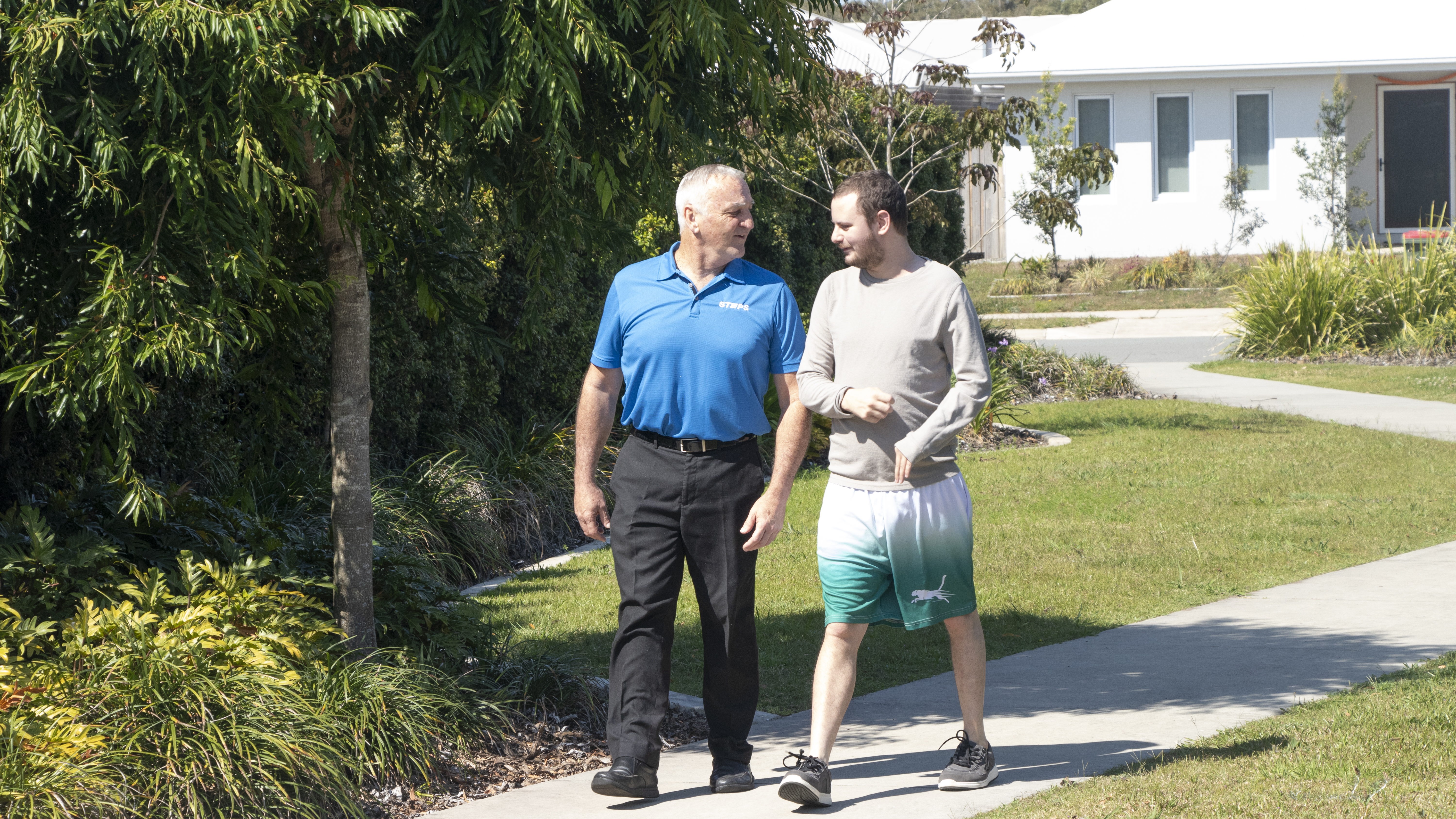 Two men walk side-by-side down a park pathway on a sunny day. The older gentleman is a STEPS Positive Behaviour Support professional; the younger man is a Positive Behaviour Support client.