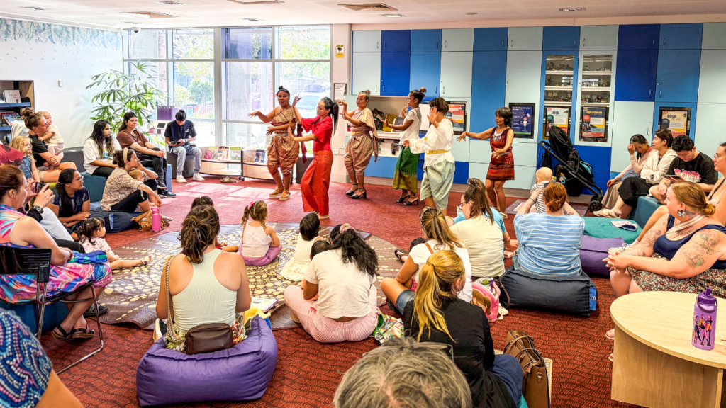 Several people sitting in a brightly-lit community space, watching a group of dancers perform.