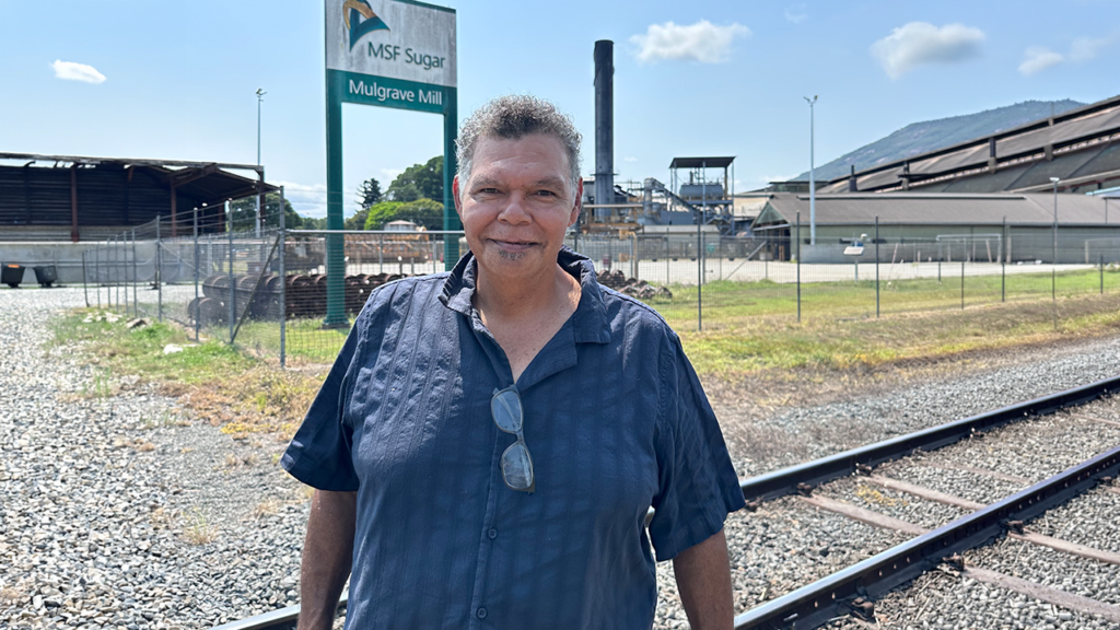 Darryl stands in front of railroad tracks. Behind him is a sign reading ‘MSF Sugar, Mulgrave Mill.’