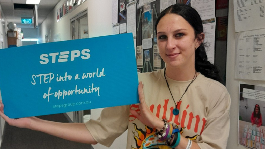 Ella-Jade stands in the hallway of the STEPS Cairns site. She is smiling and posing with a blue ‘STEPS’ poster.