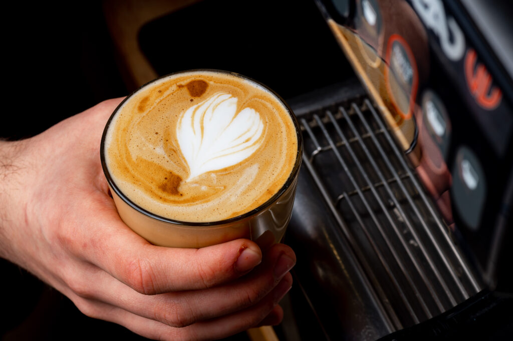 Hand holding glass of latte with heart latte art. Finished coffee beverage with white heart pattern in milk foam held by hand.