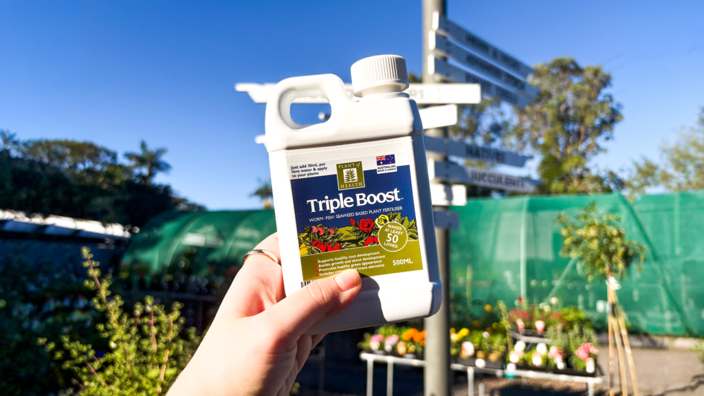 Bottle of Triple Boost plant fertiliser held in the STEPS Garden Centre with plants and flowers in the background