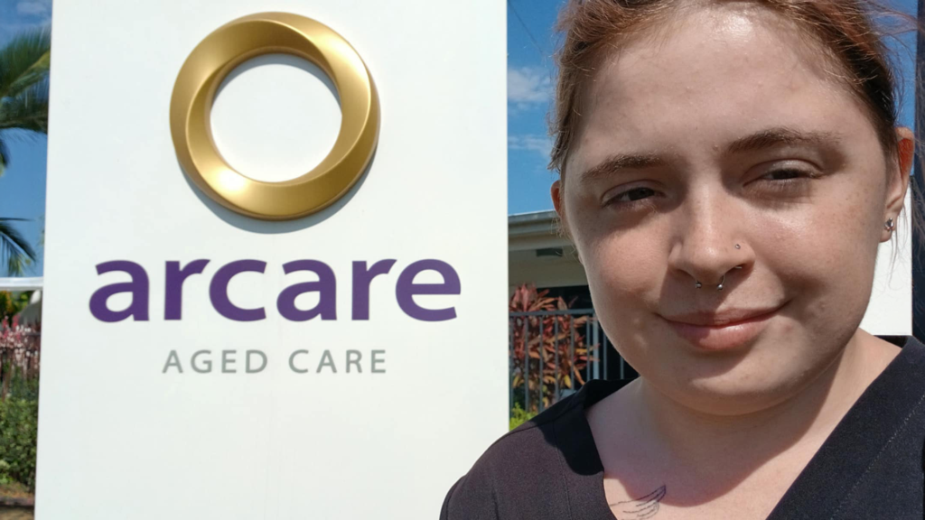 Hannah in a selfie, standing beside an Arcare Aged Care sign and smiling.