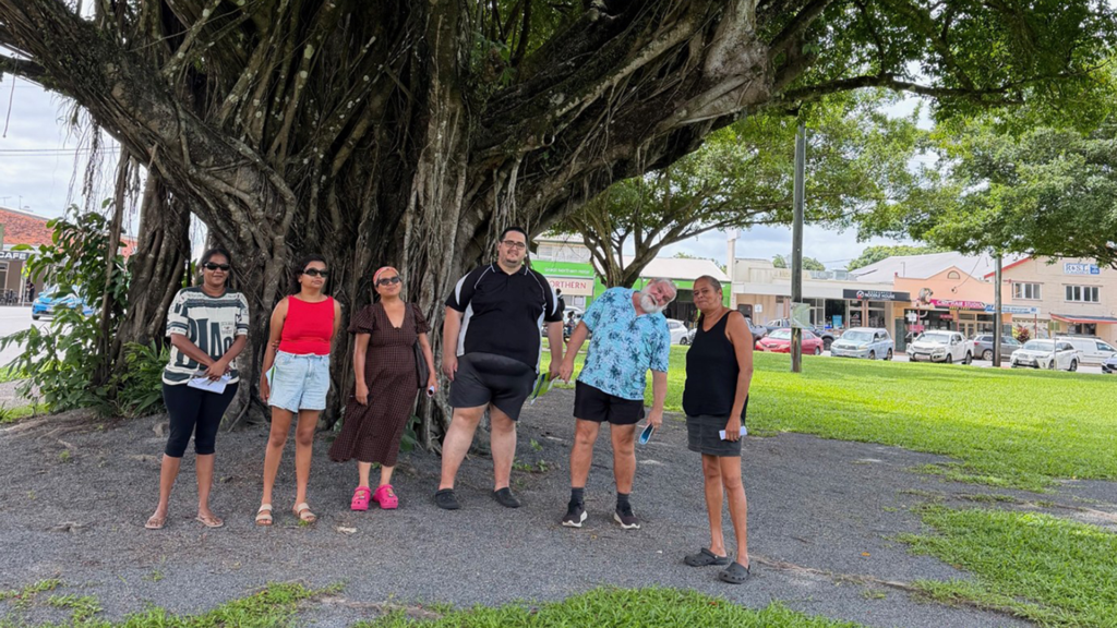 Six adults stand near the base of a large tree in a park on an overcast day, smiling at the camera.