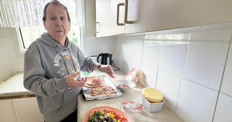 A participant prepares homemade pizzas in a kitchen, adding toppings to pizza bases on a bench alongside fresh ingredients.