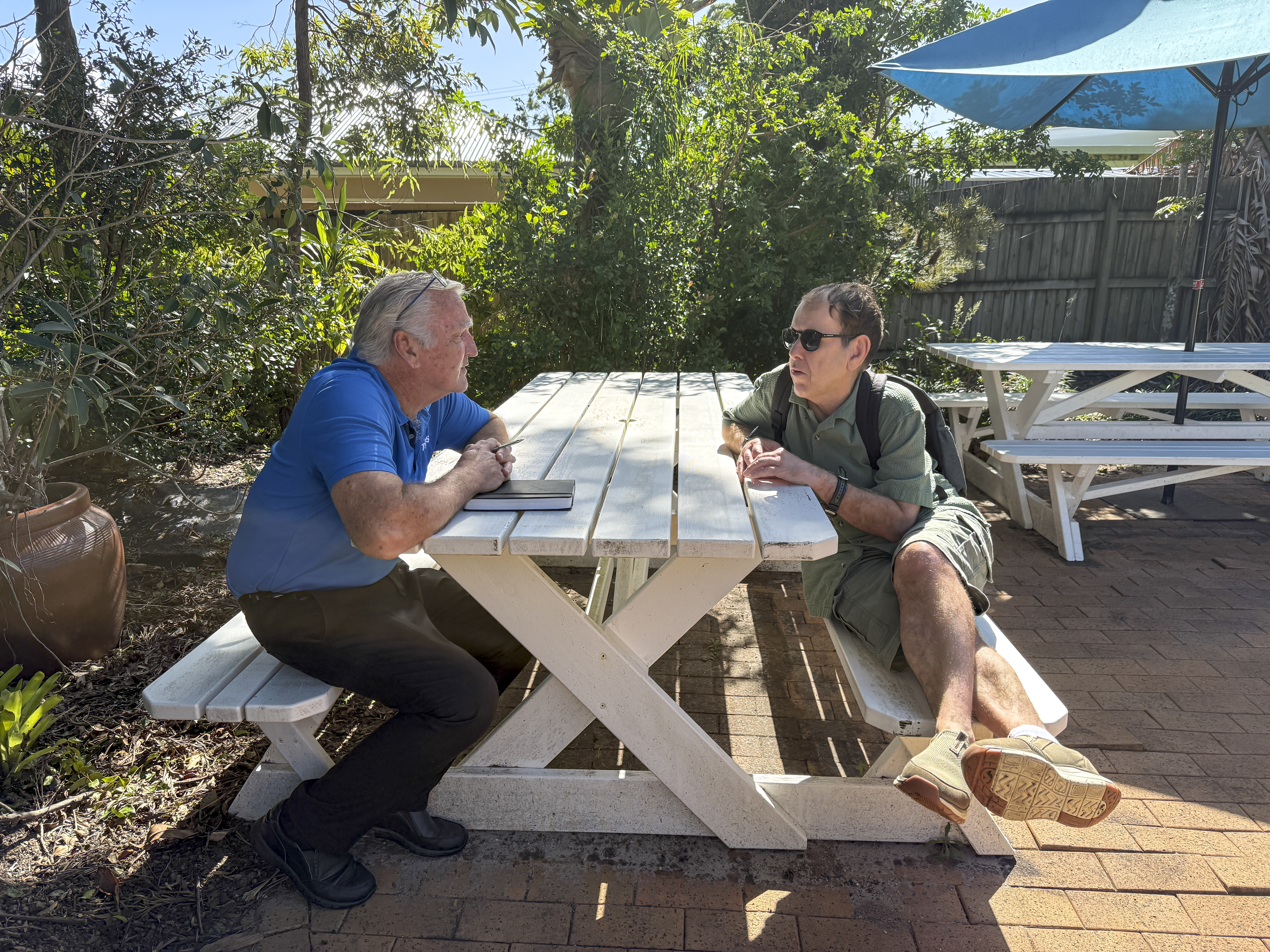 STEPS Positive Behaviour Support trainer and client sit together and talk on white bench outside, surrounded by garden on sunny day.