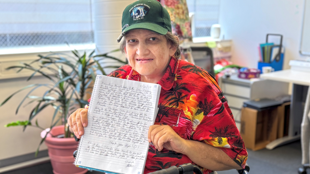 STESPS SEE Program participant Jemma sits in a classroom. She is holding a writing exercise book up to the camera, showing her journal entry about the Minister Giles visit.