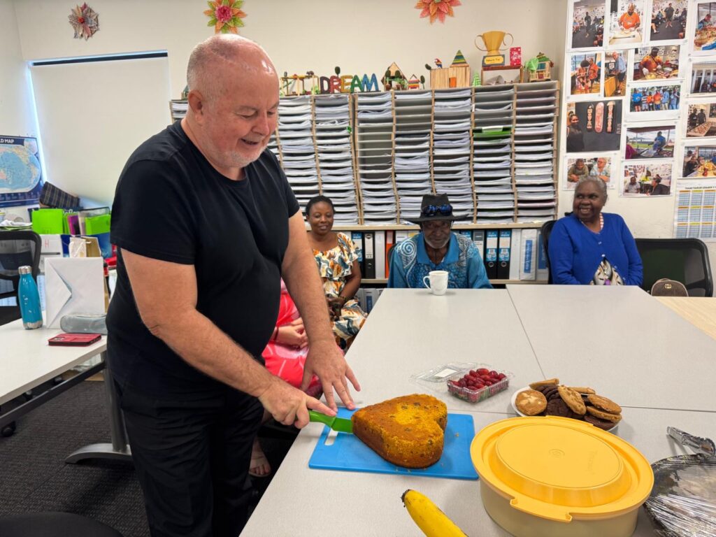 STEPS Volunteer Jurgen smiling while cutting into a heart shaped cake while SEE students are watching and smiling.