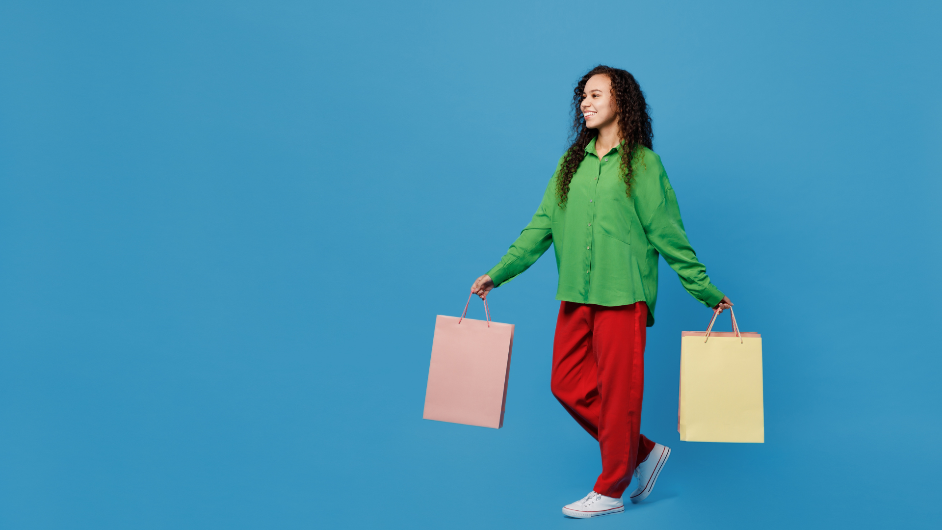 Person walking against a bright blue studio background while carrying two pastel shopping bags, wearing a green button-up shirt, red trousers and white sneakers.