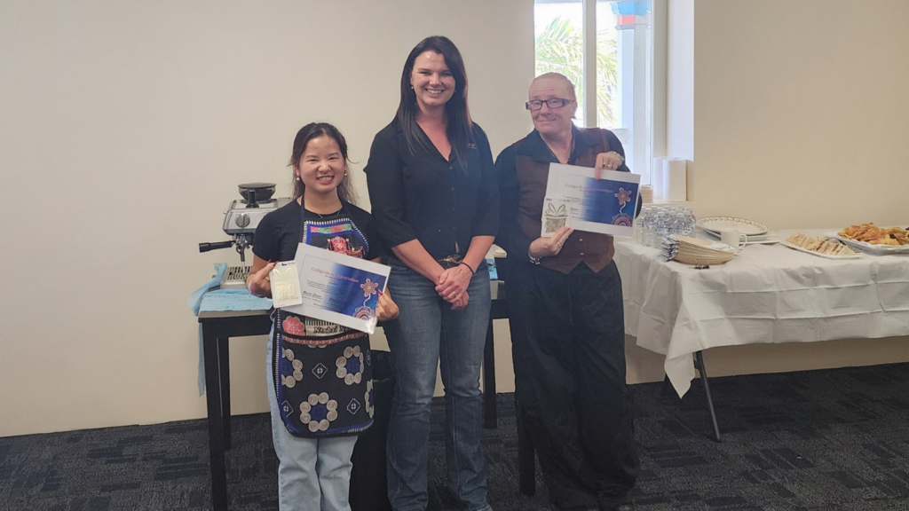 Three women smiling, standing in front of catering tables. Two of the women are holding certificates of appreciation.