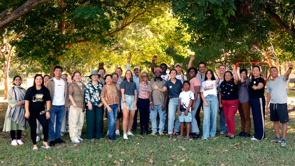 A diverse group of people standing in a park on a sunny day, smiling and waving at the camera.