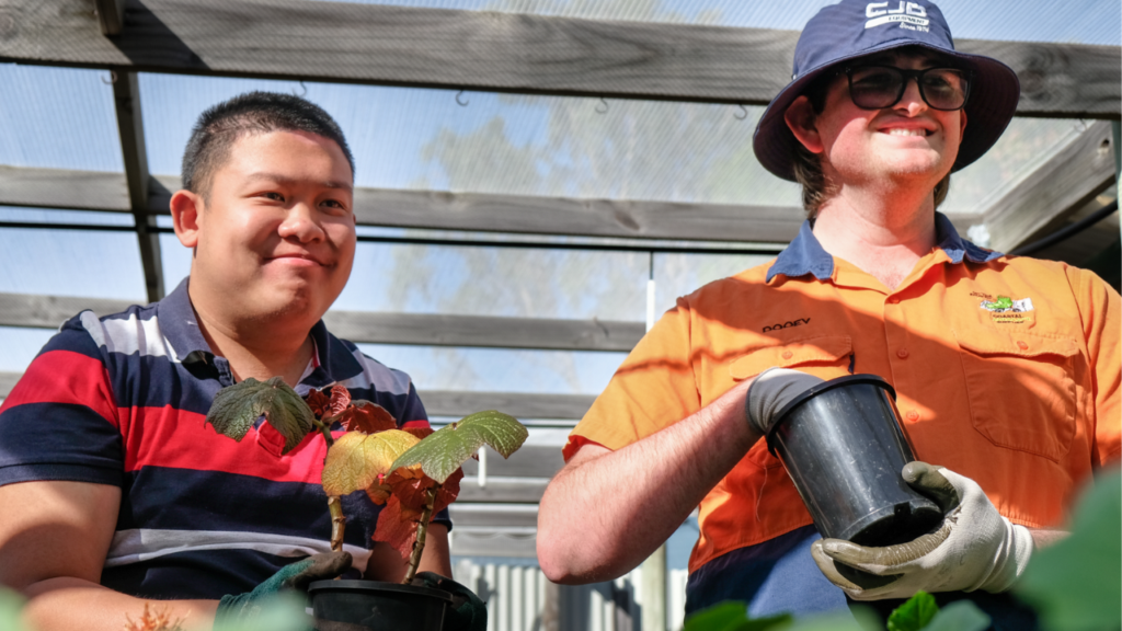 Two young men stand in a nursery, holding potted plants and smiling.