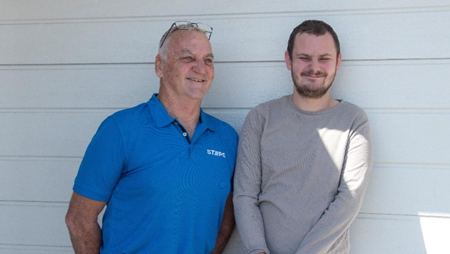  Two men stand next to one another against a white garage roller door. The older gentleman is a STEPS Positive Behaviour Support professional; the younger man is a Positive Behaviour Support client.