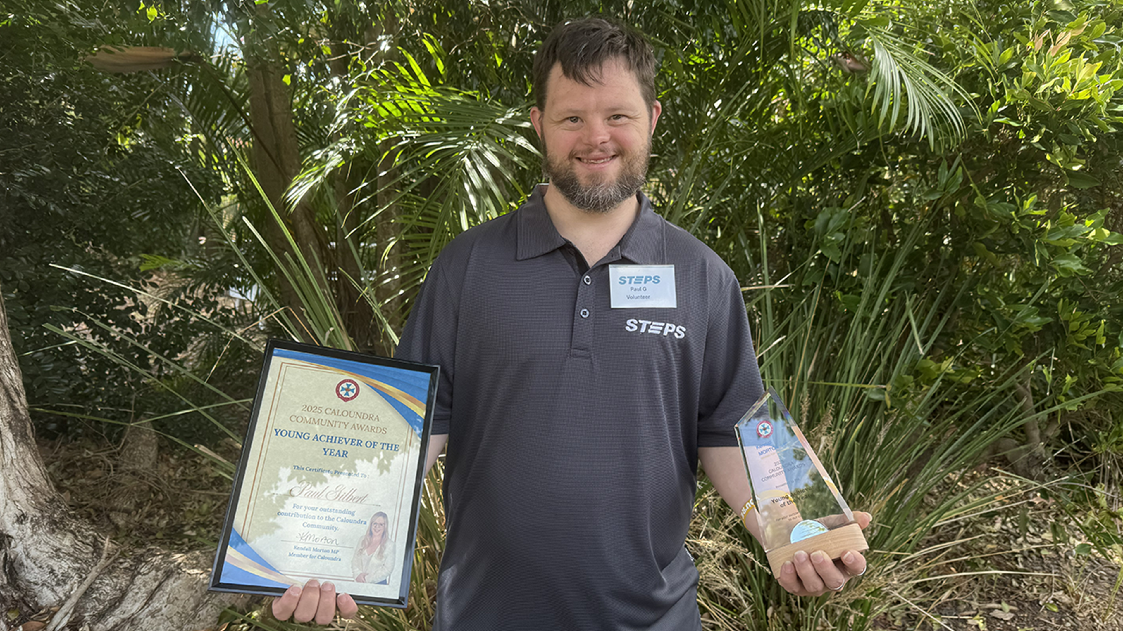 STEPS Community Support participant and volunteer Paul stands in front of lush green shrubbery. He is wearing a grey STEPS polo shirt and a name tag, and he is smiling at the camera, holding his Young Achiever of the Year trophy in his left hand and the certificate in his right hand.