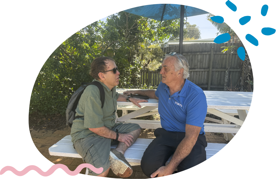 A STEPS Positive Behaviour Support practitioner sits with a participant at an outdoor table, engaging in a one-on-one conversation in a garden setting.