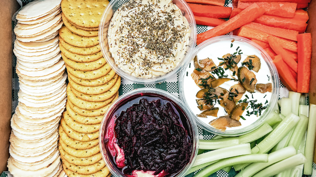 Tzatziki, basil pesto, and baba ganoush dips nestled between crackers, carrot sticks and celery sticks.