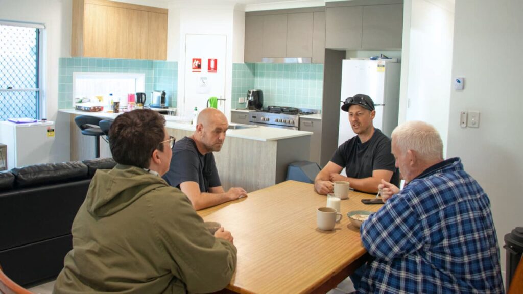 STEPS Supported Independent Living (SIL) housemates sit at dining table with Support Worker, drinking coffee and enjoying company.