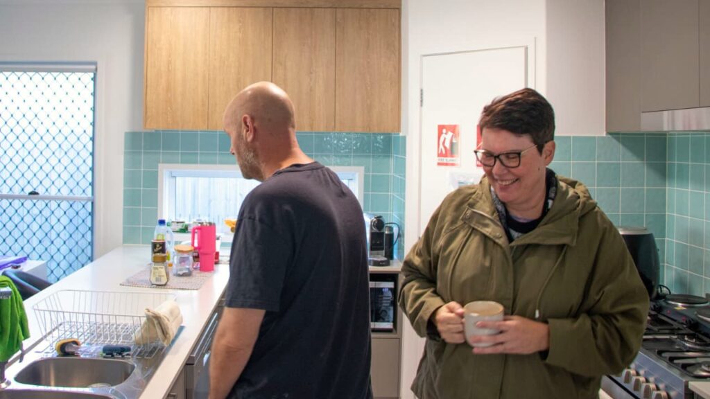 STEPS Supported Independent Living (SIL) housemates stand in kitchen; the woman is smiling and holding a cup of coffee, the man is faced toward the nearby television.