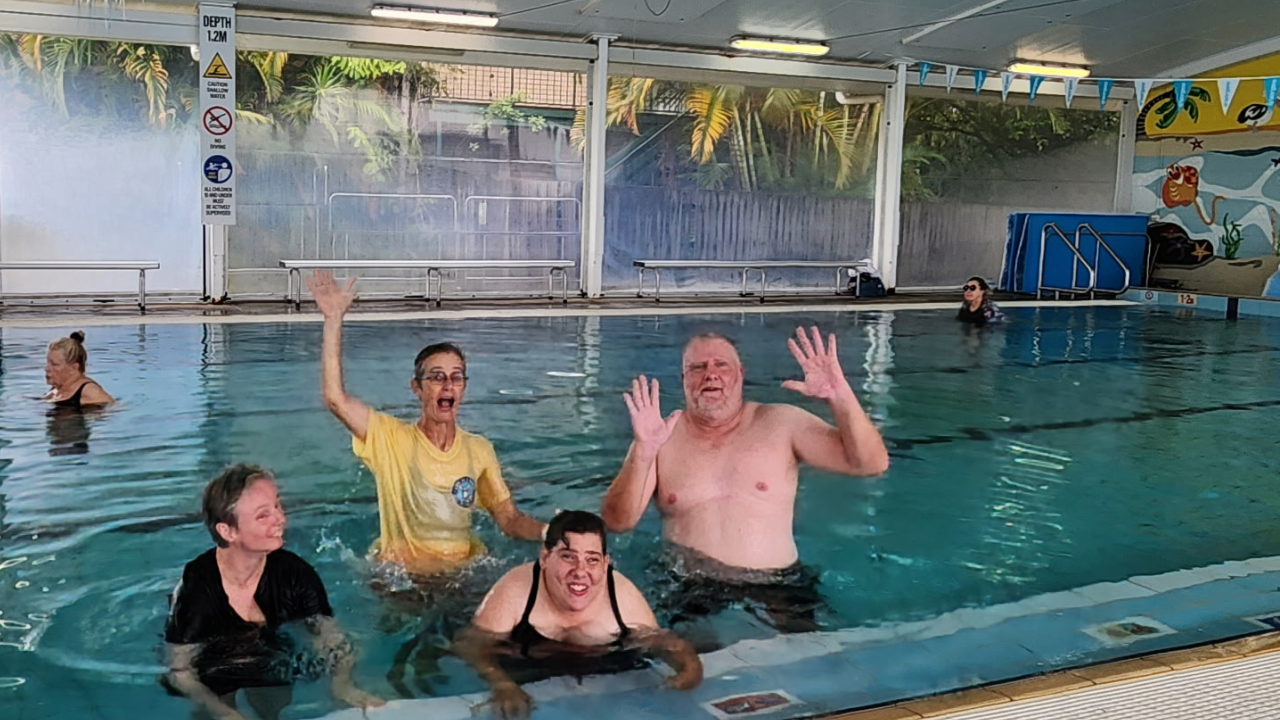 Four people are in a public undercover swimming pool, waving and smiling at camera.