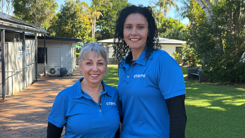 Two women stand outside on a bright, sunny day. They are wearing blue STEPS-branded polo shirts and smiling at the camera.