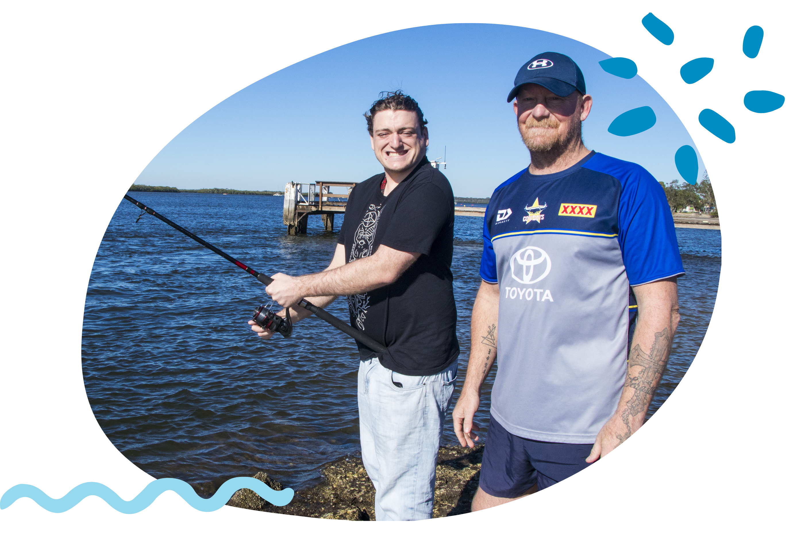 Two people stand on a rocky shoreline beside a calm body of water, with one person holding a fishing rod. A small jetty extends into the water in the background under a clear blue sky.