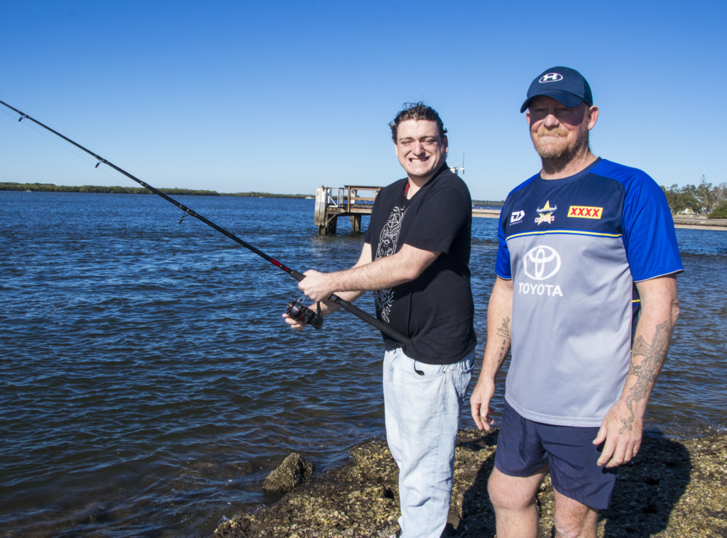 Two people stand on a rocky waterfront, with one person holding a fishing rod and reel beside a calm body of water. A small jetty extends into the water under a clear blue sky.