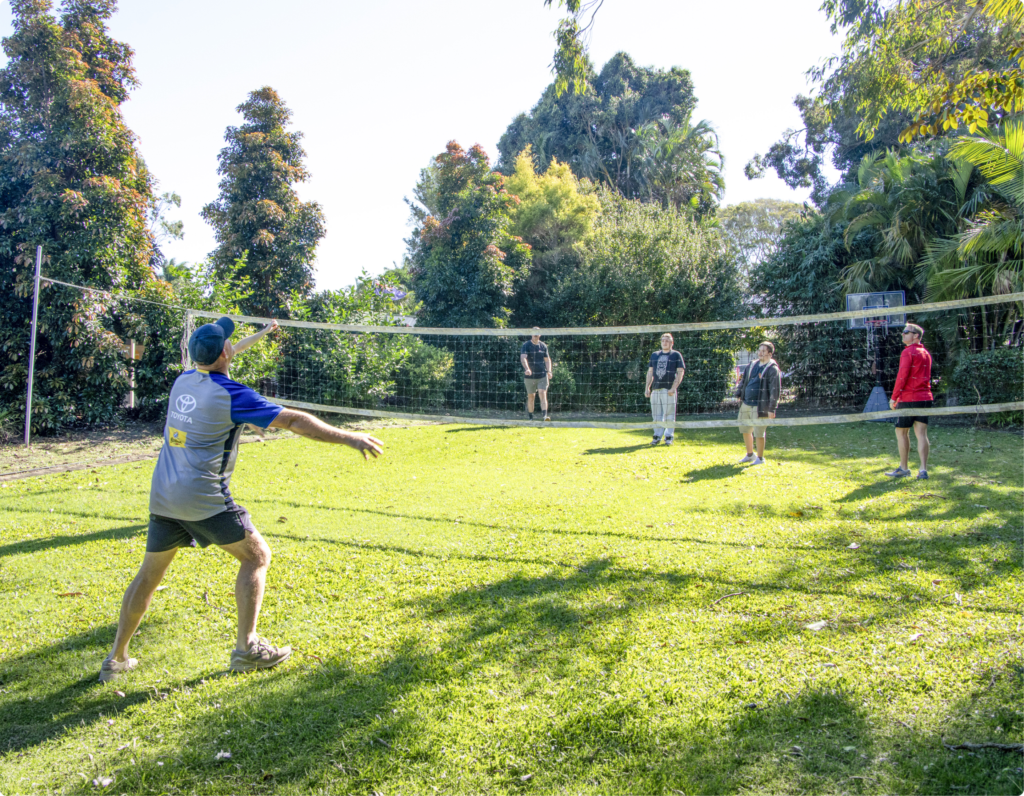 A group of people participate in an outdoor volleyball game on a grassy lawn surrounded by trees and garden vegetation
