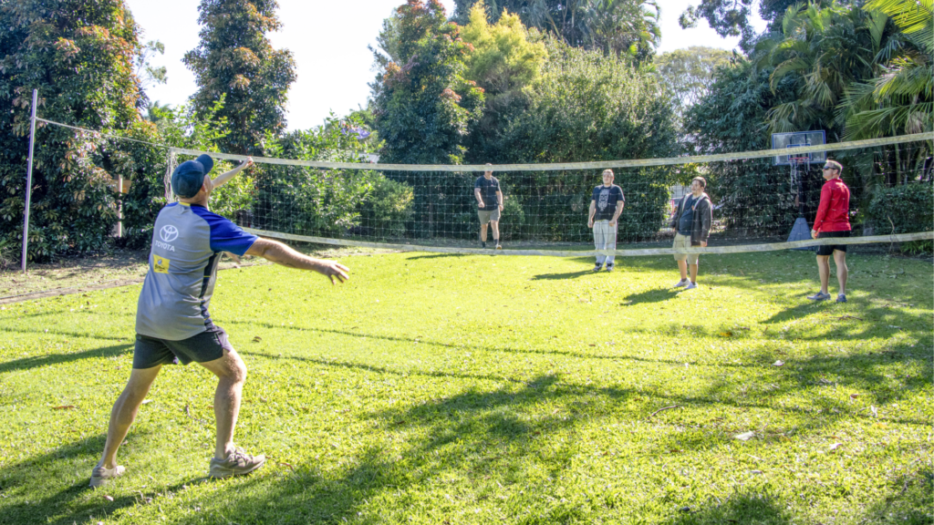 A group of people participate in an outdoor volleyball game on a grassy lawn surrounded by trees and garden vegetation