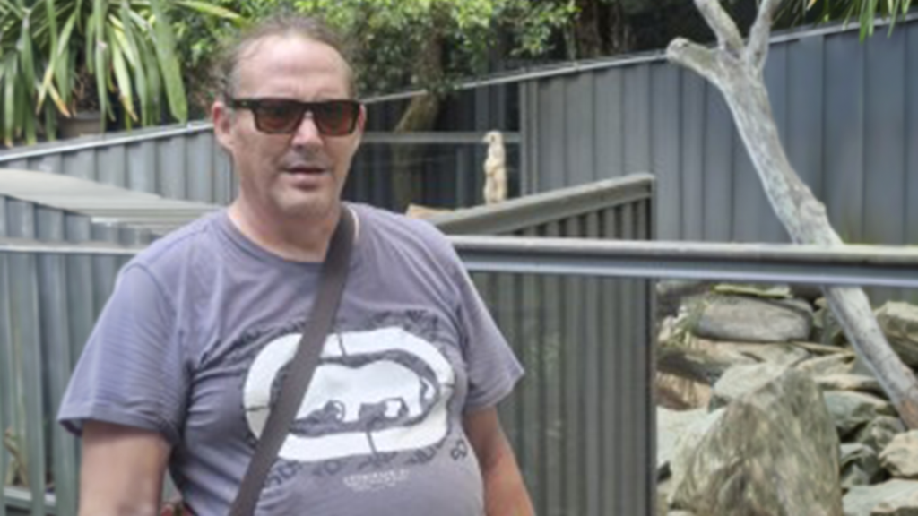 Four people are in a public undercover swimming pool, waving and smiling at camera.