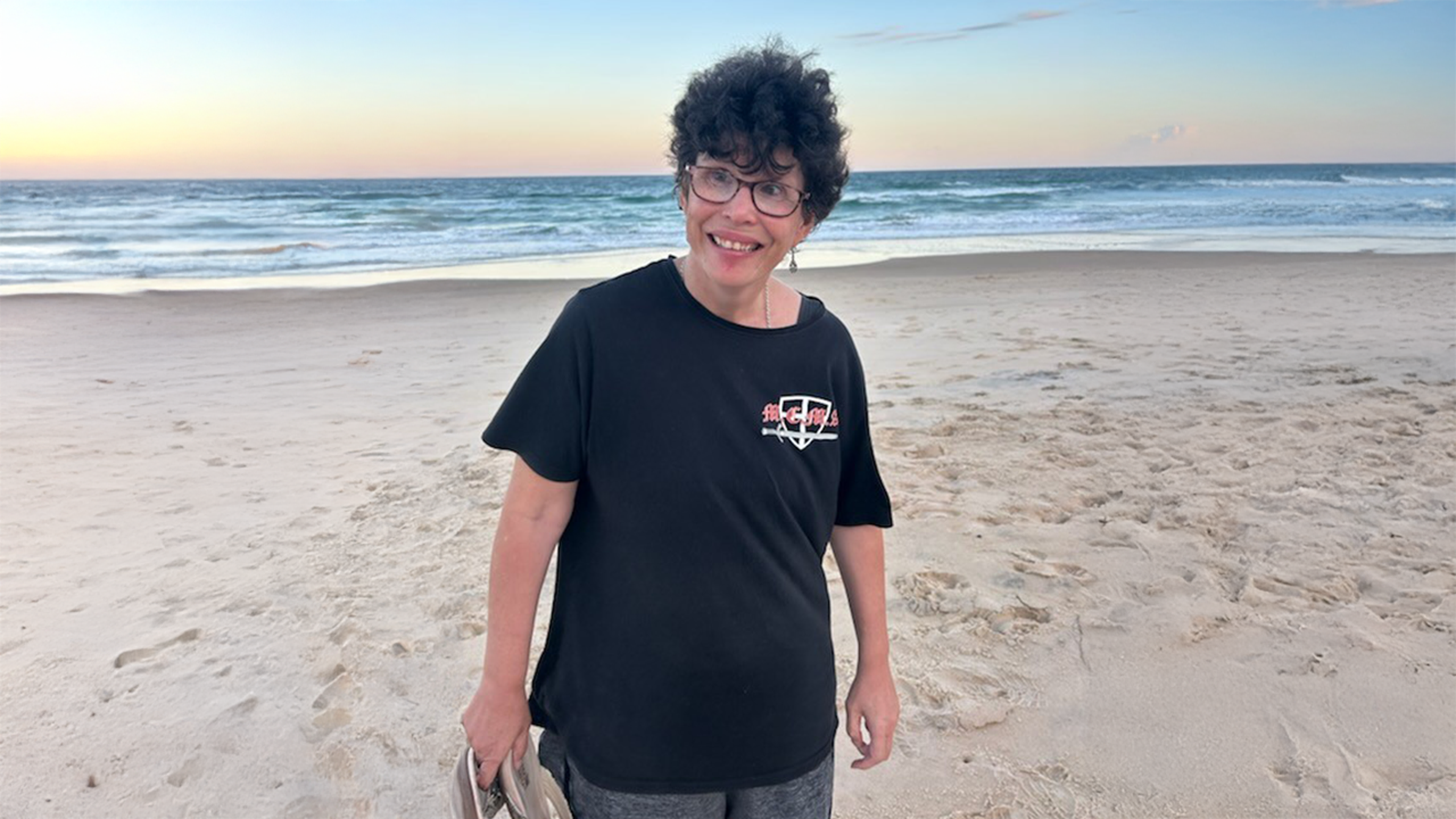 STEPS Community Support participant Sarah stands on a Gold Coast beach, with the white sand, ocean and morning sky behind her. She is holding her shoes in one hand and smiling at someone slightly off-camera. She is wearing a black t-shirt and grey board shorts, and very dark brown hair and glasses.