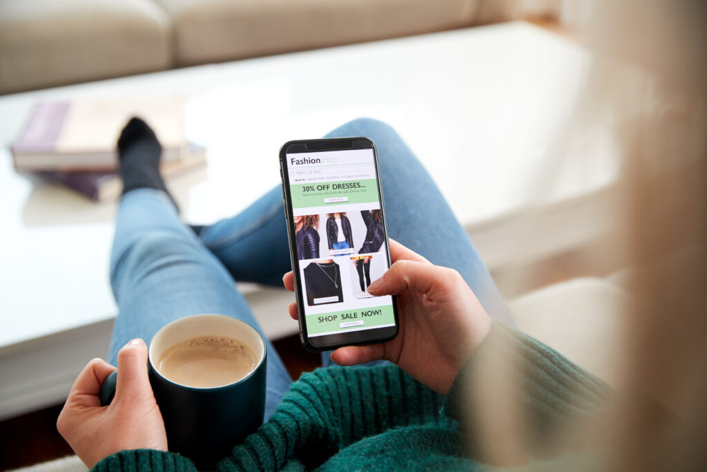 Person relaxing with a cup of coffee while browsing a fashion sale on a smartphone, showing discounted clothing items.