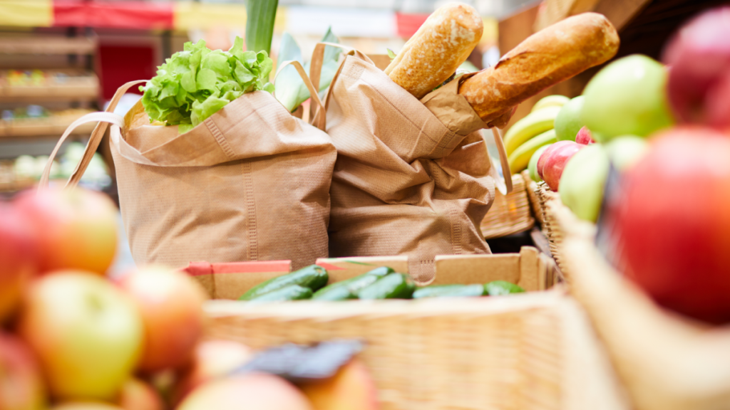 Two paper grocery bags filled with fresh produce and baguettes sit in a fruit and vegetable aisle, with apples and cucumbers visible in the foreground.