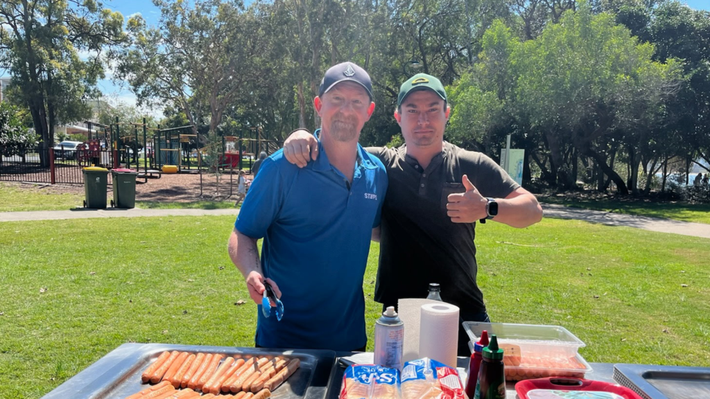 Two men stand behind public BBQ in a park on a sunny day, smiling at the camera. They have their arms around each other's shoulders, and the man on the right is giving a thumbs-up.