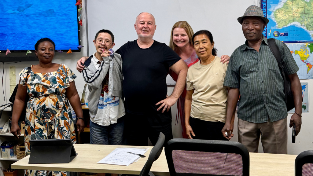 STEPS volunteer Jurgen stands in a colourful classroom in front of a whiteboard. He is surrounded by five STEPS SEE Program participants, and all are smiling at the camera.