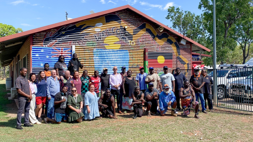 The Hon Andrew Giles MP, Minister for Skills and Training stands with a crowd of STEPS SEE Program participants and trainers at Venndale Rehabilitation Centre. They are gathered in front of a shed painted with a mural that blends Australian and First Nations culture. It is a bright and sunny day.