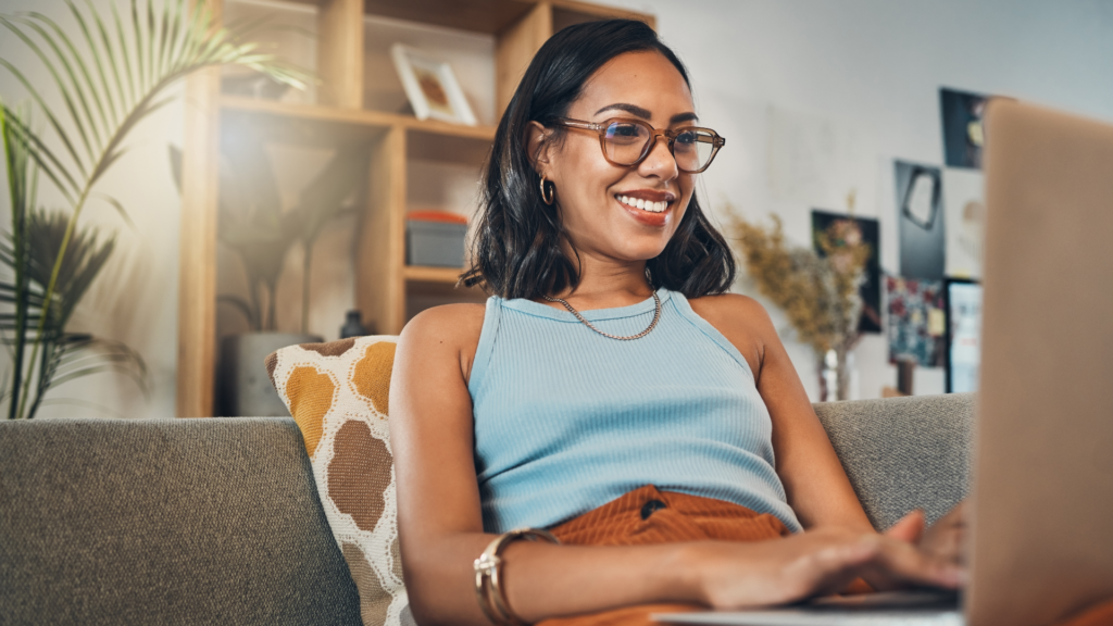 A woman sitting on her lounge at home, using her laptop. She is wearing glasses and smiling.