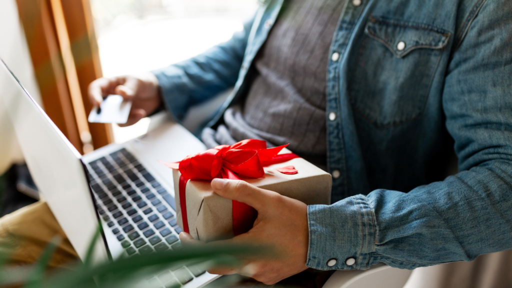 A person holding a small gift wrapped in brown paper with a red ribbon while using a credit card next to a laptop.