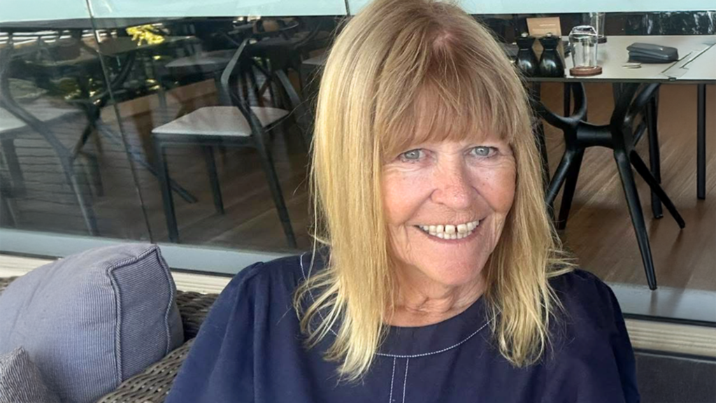 Yvonne Coleman, a blonde woman with a full fringe, smiles at the camera. It is a close-up photo. She is sitting on an outdoor lounge at a restaurant in the daytime and wearing a dark blue shirt.