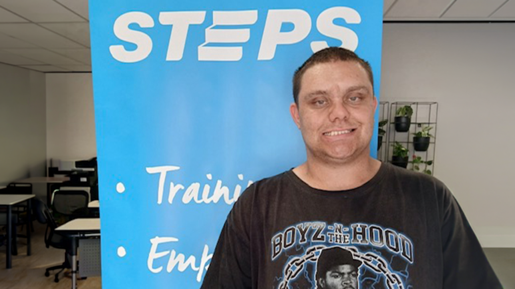 Zak stands in front of a bright blue STEPS banner, which lists ‘Training, Employment, Support, NDIS.’